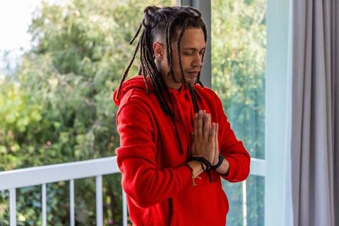 Man practicing meditation on balcony wearing red hoodie and dreadlocks in morning sunlight