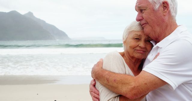 Elderly couple embracing on picturesque beach with mountain in background. Ideal for themes related to senior love, retirement, vacation, outdoor activities, or the beauty of nature. Perfect for promoting retirement communities, travel destinations, lifestyle blogs, or senior health and wellness.