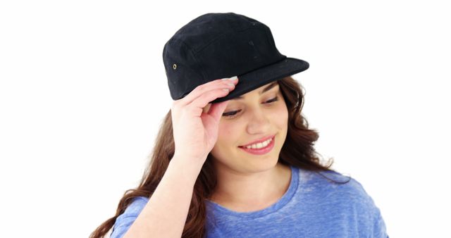 Young woman smiling while adjusting her black cap, wearing a casual blue shirt. Close-up shot on white background. Ideal for use in fashion blogs, lifestyle magazines, casual wear advertisements, or social media profiles showcasing casual and modern styles. Promotes a relaxed, youthful, and approachable vibe.
