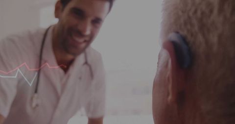 Doctor leaning and consulting senior patient wearing hearing aid in backlit clinic