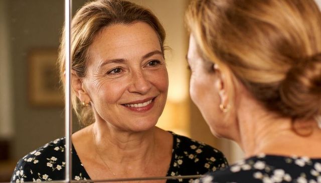 Smiling middle-aged woman checking mirror reflection on home vanity, warm natural light