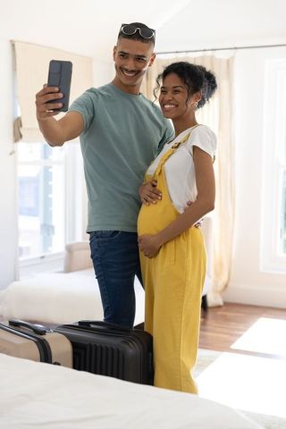 Expectant couple taking selfie in sunlit bedroom while cradling baby bump near packed luggage