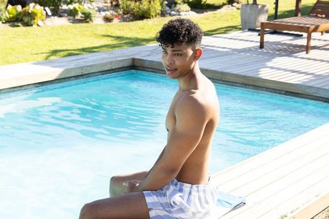 Young man relaxing on pool deck wearing blue striped trunks on sunny day