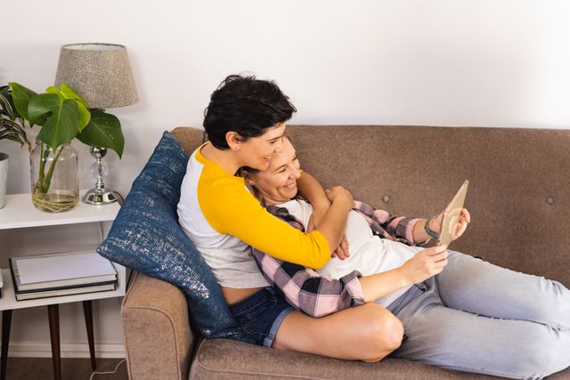 Lesbian couple relaxing on sofa, enjoying video on tablet. Perfect for use in articles about LGBTQ+ relationships, home lifestyle, technology in daily life, and modern love. Ideal for promoting inclusivity, digital devices, and domestic happiness.
