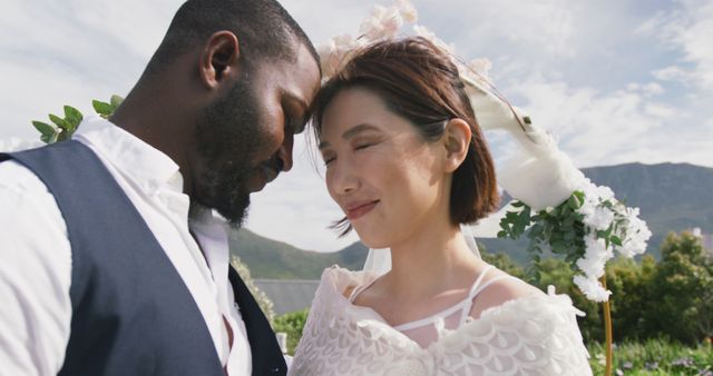 Image of happy diverse bride and groom touching heads together and smiling at outdoor wedding. Marriage, love, happiness and inclusivity concept.