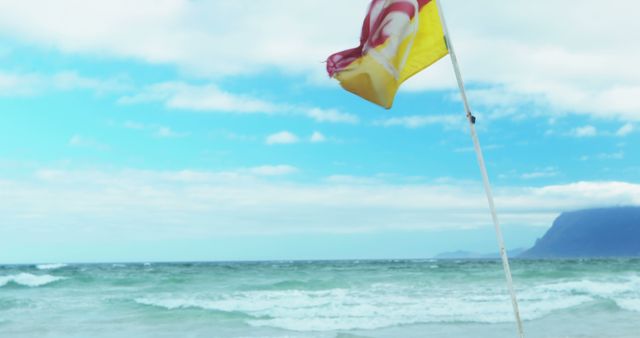 Yellow flag on a sandy beach fluttering in the wind with ocean waves in the background. Mountain range and partly cloudy sky add to the scenic beauty. Ideal for travel and vacation websites, safety warnings, and outdoor activity promotions.