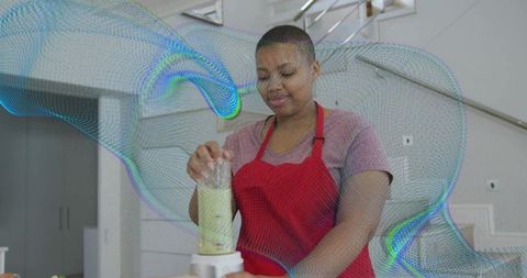 Young woman blending green smoothie in modern kitchen wearing red apron, healthy lifestyle