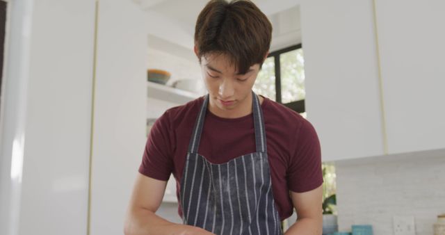 A young Asian man is wearing an apron and engaging in cooking in the kitchen. The kitchen has a bright and clean style, with white cabinets and natural lighting. This image can be used for content related to cooking tutorials, lifestyle blogs, home cooking promotion, kitchen appliance advertisements, or culinary education.