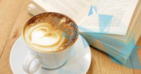 Sitting white ceramic cappuccino cup with heart latte art beside open book on wooden table