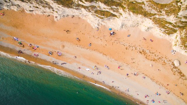 This image showcases an aerial view of a sandy beach lined with a coastal cliff. Visitors are seen relaxing and sunbathing, capturing a typical summer vacation day. It is ideal for travel brochures, posters, tourism websites, and articles related to beach vacations, outdoor activities, and nature retreats.