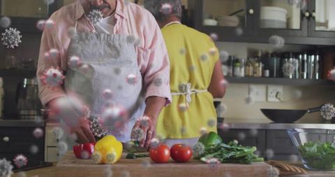 Senior couple cooking at home wearing aprons slicing bell peppers with virus overlay