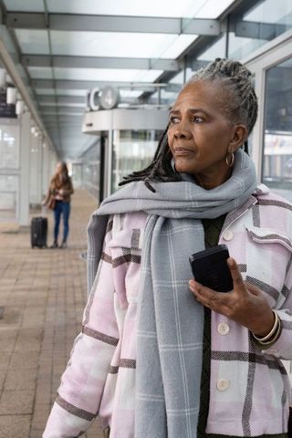 Senior black woman tapping smartphone on urban transit platform waiting for train