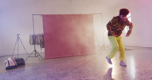 Fashion designer wearing vibrant clothes dancing in a stylish loft studio during a photo shoot. Photo includes photographer's studio setup with clothing rack, tripod, and backdrop. This image is ideal for illustrating creativity in fashion, behind-the-scenes of studio work, artistic environments, and promotional content for fashion brands.