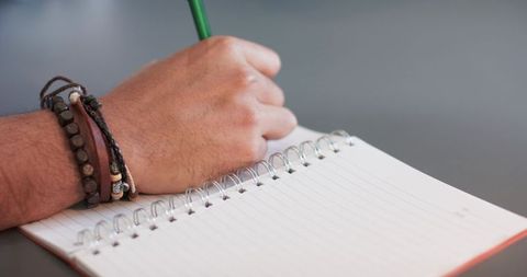 Man writing in spiral notebook wearing layered leather and bead bracelets at desk