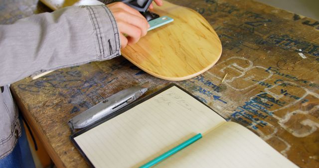 A person is sanding a wooden circle on a workbench covered in scribbles, with a notebook and pencil nearby, indicating a planning or creative process. Capturing a moment of craftsmanship, the image suggests a DIY project or woodworking activity.