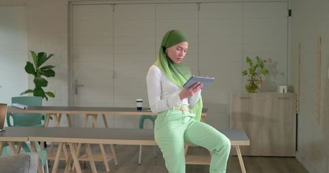 Asian muslim woman wearing green hijab using tablet in minimalist coworking workspace