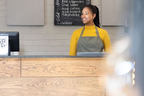 Smiling african american barista wearing mustard top and olive apron behind coffee counter
