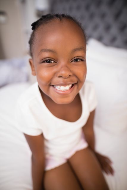 This image captures a joyful young girl sitting on a bed at home, smiling brightly. Ideal for use in family-oriented content, advertisements for children's products, lifestyle blogs, and educational materials. The warm and inviting atmosphere makes it suitable for promoting home decor, parenting tips, and health and wellness topics.