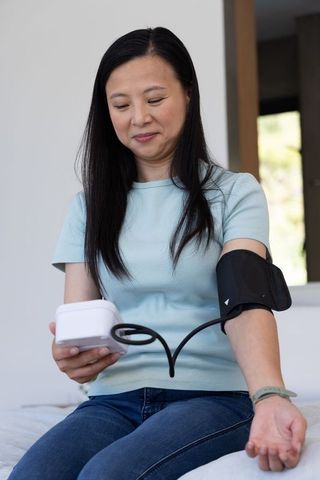 Mature asian woman checking blood pressure at home with white digital cuff and monitor