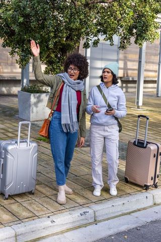 Female friends traveling standing on curb waving while waiting with rolling suitcases