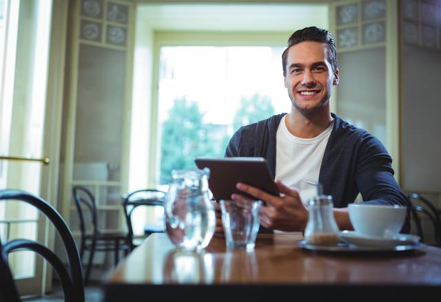Young man sitting in a cozy café, smiling while using a digital tablet. Ideal for themes related to modern lifestyle, technology use in daily life, urban living, and leisure activities. Suitable for marketing materials, blog posts, and advertisements focusing on digital connectivity, casual dining, and relaxed environments.