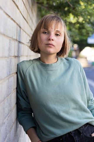 Young woman leaning against brick wall wearing green crewneck and nose stud
