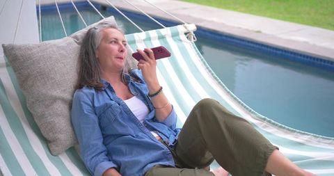 Middle-aged woman relaxing in striped hammock by pool speaking into smartphone on speaker
