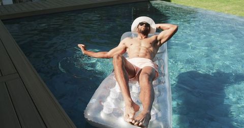 Tanned man relaxing on clear inflatable float in backyard pool wearing sun hat and sunglasses