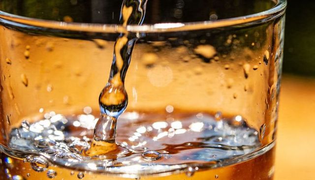 Macro water stream pouring into glass capturing suspended droplet, ripples and warm reflections