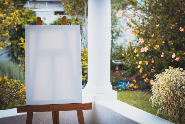 Blank canvas on an easel placed on a terrace, surrounded by a vibrant summer garden. Ideal for themes related to creative hobbies, home quarantine activities, outdoor painting, and artistic inspiration. Can be used for promoting art supplies, encouraging creativity, or illustrating peaceful home environments.