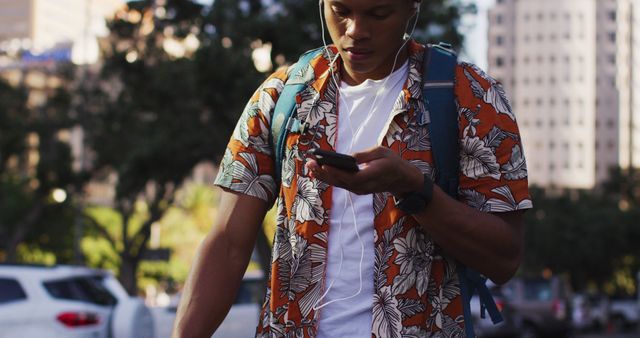 The image features a young man wearing a brightly patterned shirt, walking in an urban environment with skyscrapers and greenery. He is listening to music with earphones and checking his phone, suggesting a digitally connected lifestyle. Ideal for illustrating urban life, technology use, or transportation themes in advertisements, articles, or social media.