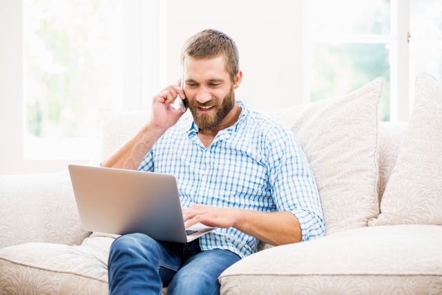 Man using laptop while talking on mobile phone in living room at home