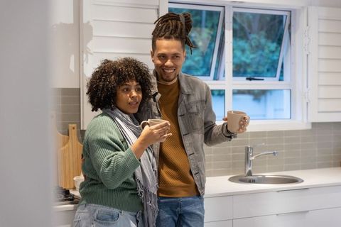 African american couple standing in cozy kitchen holding mugs, smiling over morning coffee