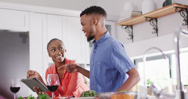 An African American couple is spending quality time in the kitchen preparing a meal together. The man is holding a tablet, likely looking up a recipe, while the woman smiles at him. They have vegetables and wine on the countertop, indicating a casual and bonding moment. This image can be used in articles, blogs, or advertisements focused on relationships, cooking, lifestyle, home living, or kitchen activities.