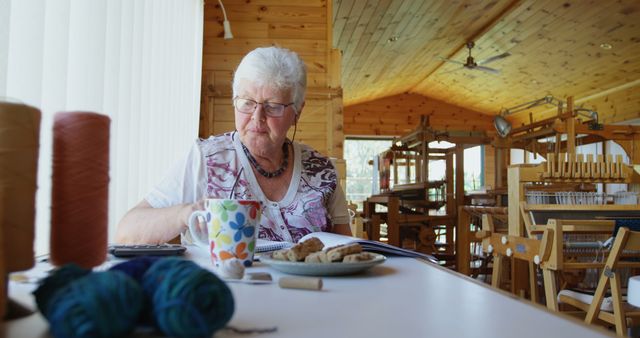Senior Caucasian woman enjoys a break in a craft room, with copy space. She's surrounded by yarn and weaving looms, indicating a creative hobby setting.