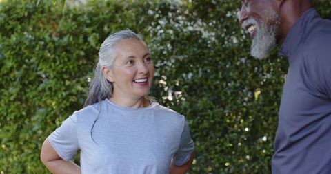Mature diverse couple laughing and chatting outdoors in workout clothes by green hedge