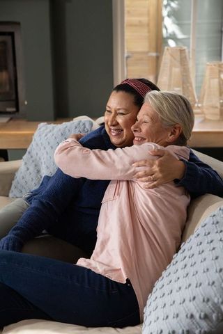 Diverse women hugging on cozy living room sofa, smiling friends sharing warm home embrace