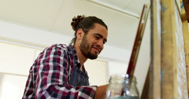 Young bearded man in plaid shirt painting at easel in a bright art studio. Image ideal for articles on creativity, self-expression, artist profiles, or educational content about painting.
