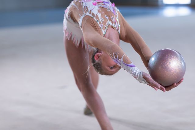 Caucasian female gymnast practicing at the gym, stretching backwards and holding a pink ball in her hands. Gymnast training hard for competition.