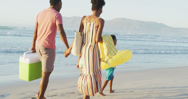 Family walking along beach holding cooler and float, enjoying summer day. Perfect for travel and tourism advertising, vacation brochures, family lifestyle blogs, and magazines promoting outdoor activities.