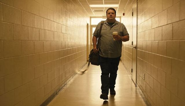 Commuter walking down narrow institutional corridor holding coffee cup and satchel