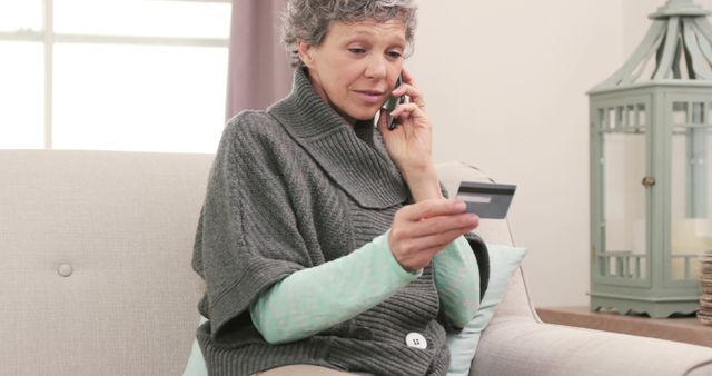 Senior woman sitting on sofa using her phone and credit card for online shopping. Ideal for concepts related to senior citizens handling finances, technology use among older adults, or comfortable home living.