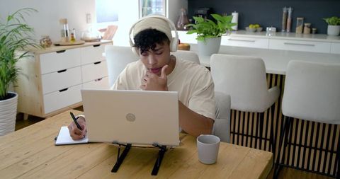 African american man studying at home wearing headphones and taking notes on laptop