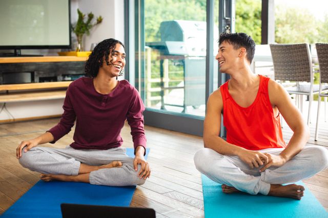 Multiracial gay couple looking at each other and laughing while meditating on mats at home. Happy, unaltered, love, together, homosexual, exercise, yoga, fitness and home concept.