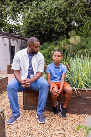 African american dad and teen son sitting and talking on backyard planter wearing sneakers