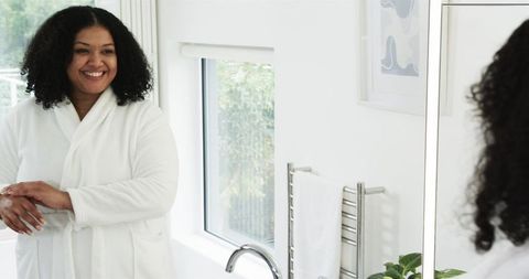 Mid adult African American woman wearing white bathrobe smiling at mirror in bathroom