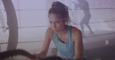 Determined woman gripping battle ropes in boxing ring with shadow boxer shadowboxing behind