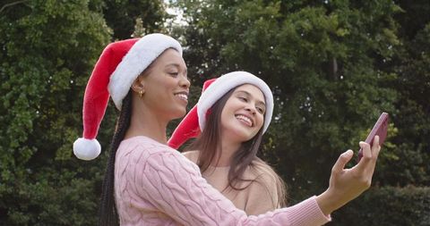 Friends taking festive selfie wearing santa hats and knit sweaters smiling in garden