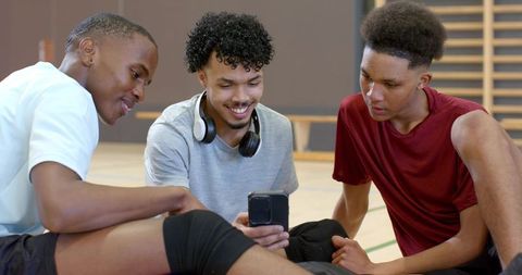 Young black teammates sharing smartphone and smiling on indoor basketball court