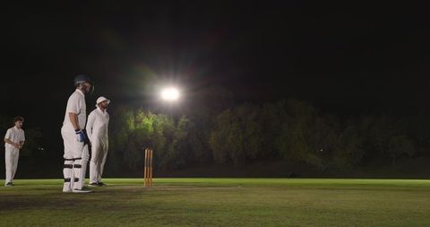 Night cricket players standing at crease under bright floodlights on pitch, dramatic